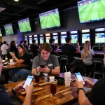 Pelanggan yang antusias menggunakan SPBO untuk taruhan olahraga langsung dalam suasana bar yang hidup.